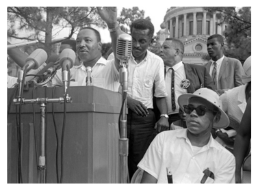 Dr. Martin Luther King speaks at the Mississippi State Capitol; James Meredith sits beside him.