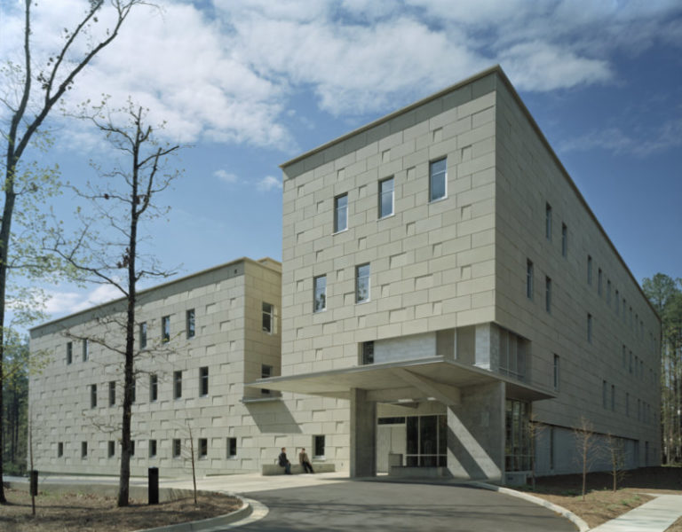 Exterior view of the Mississippi Library Commission building featuring a modern three-story stone facade, large entrance canopy, and surrounding trees under a partly cloudy sky.