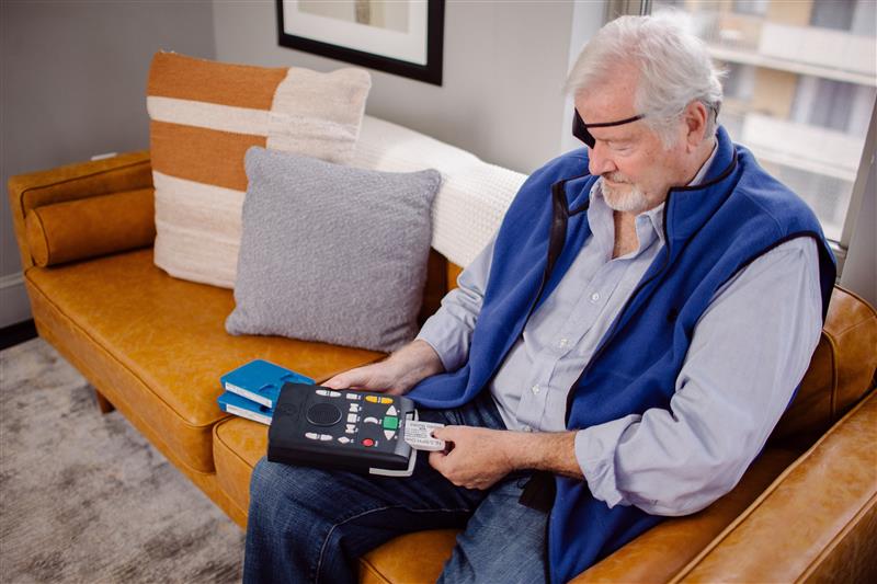 Man__Eye_Patch__DTBM_insert Visually impaired patron sits on a couch and inserts a cartridge into a digital talking book machine.