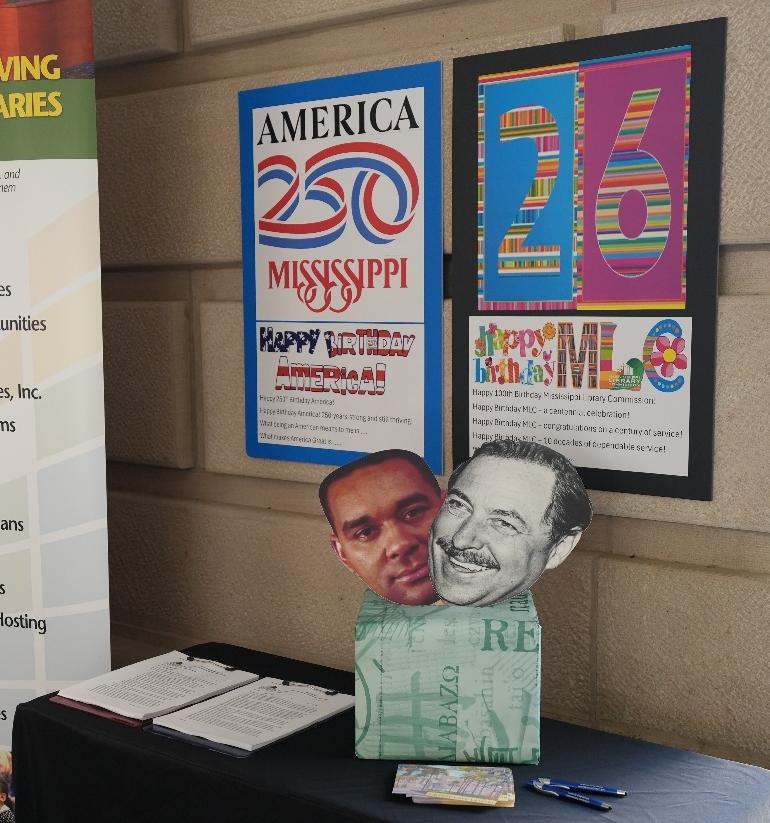 America 250 and MLC Posters Table with notebooks and pens in front of posters reading ‘America 250 Mississippi’ and ‘Happy Birthday MLC 26’ on a stone wall.