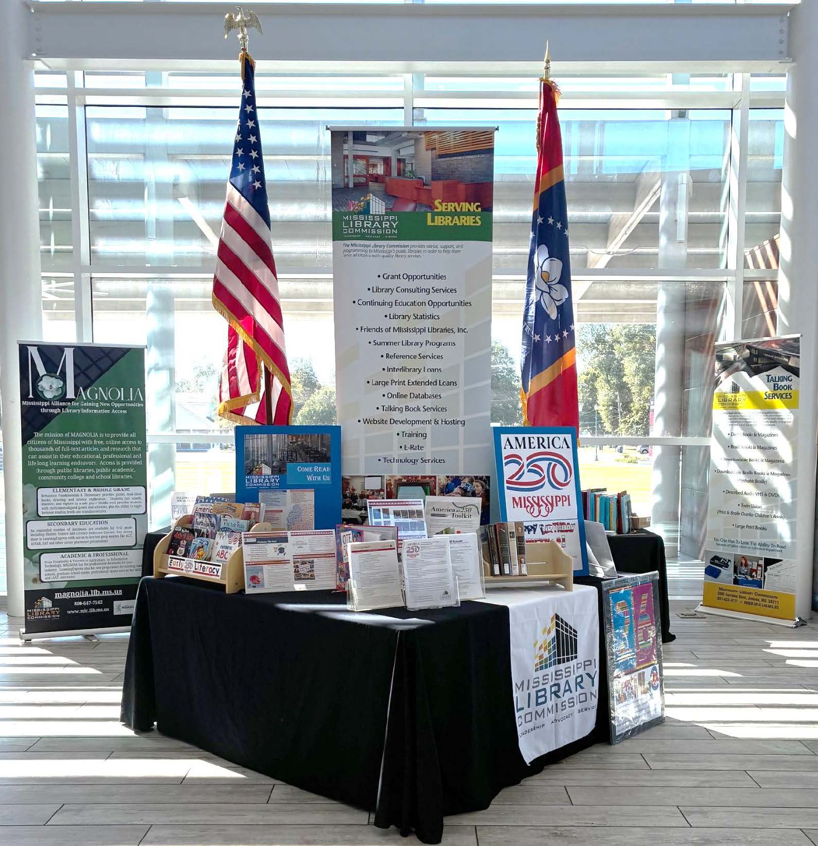Bright-AM250-Booth Mississippi Library Commission booth with America 250 signage, U.S. and state flags, books, and informational banners displayed in a bright glass-walled space.