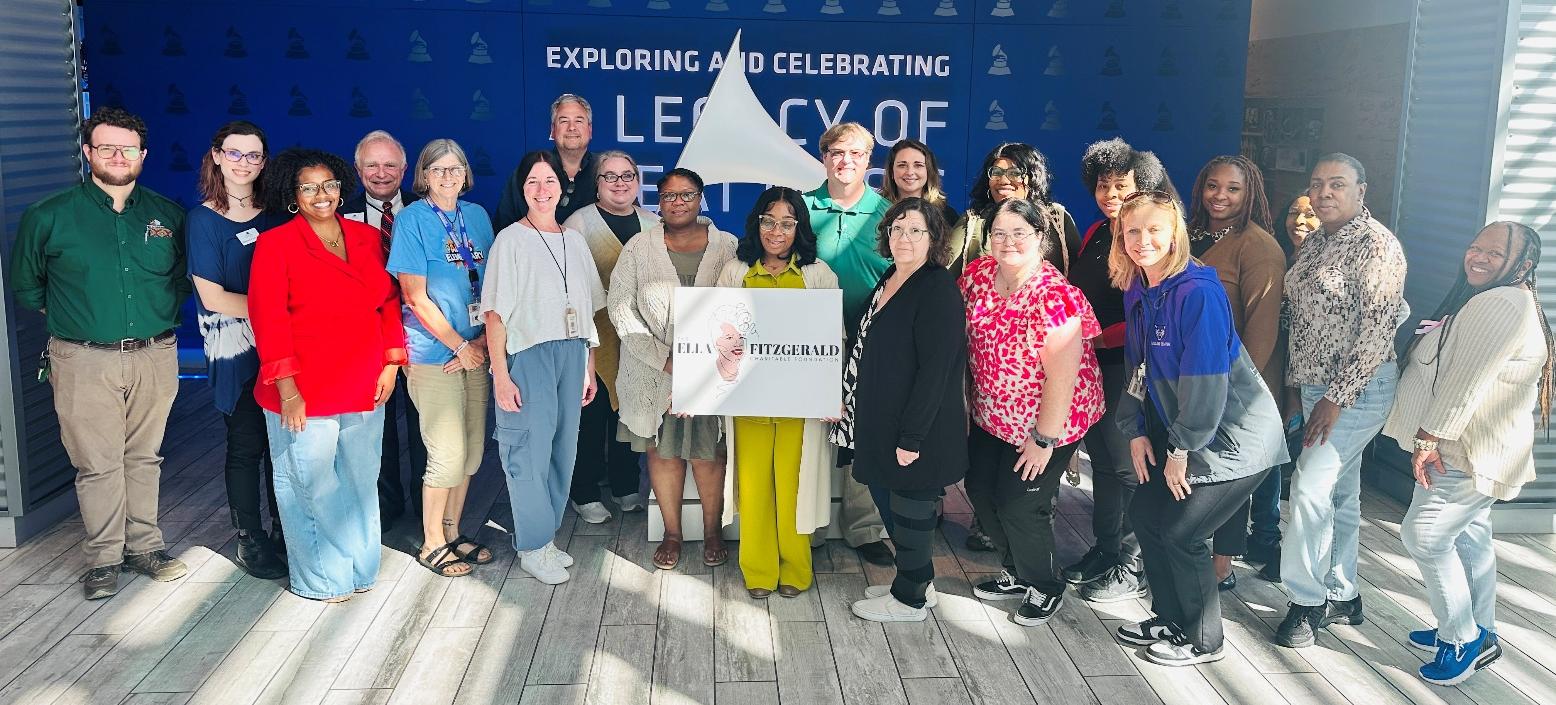 Bright AM250 Booth Group of people standing together indoors in front of a blue backdrop with text about exploring and celebrating legacy, holding a sign that reads ‘Ella Fitzgerald.’