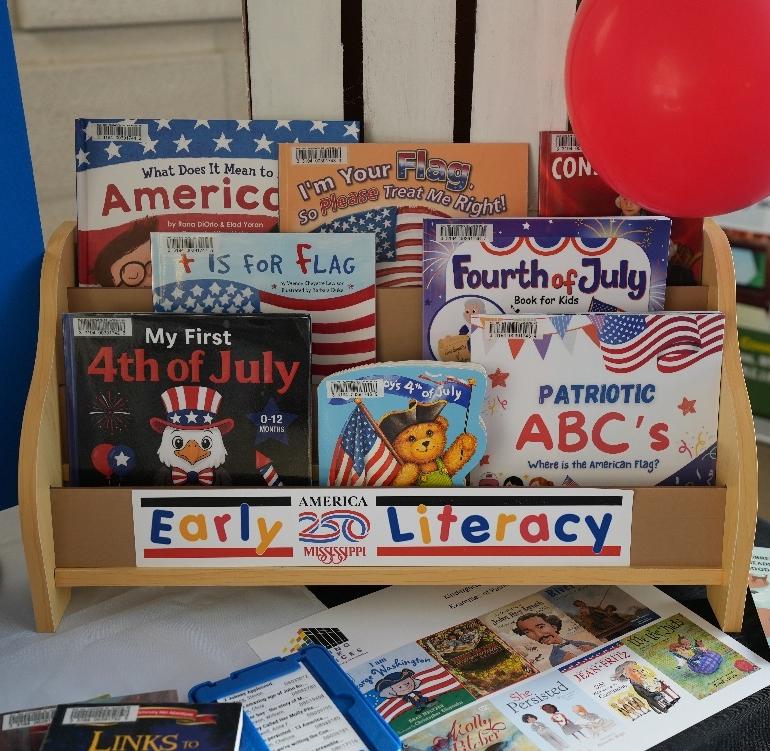 Early Literacy Book Display Wooden display rack labeled ‘Early Literacy’ holding children’s books about America, flags, and the Fourth of July, with a red balloon on the side.