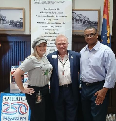 Group Photo in front of MLC Booth Three people standing in front of a Mississippi Library Commission booth with an America 250 poster and a banner listing library services.