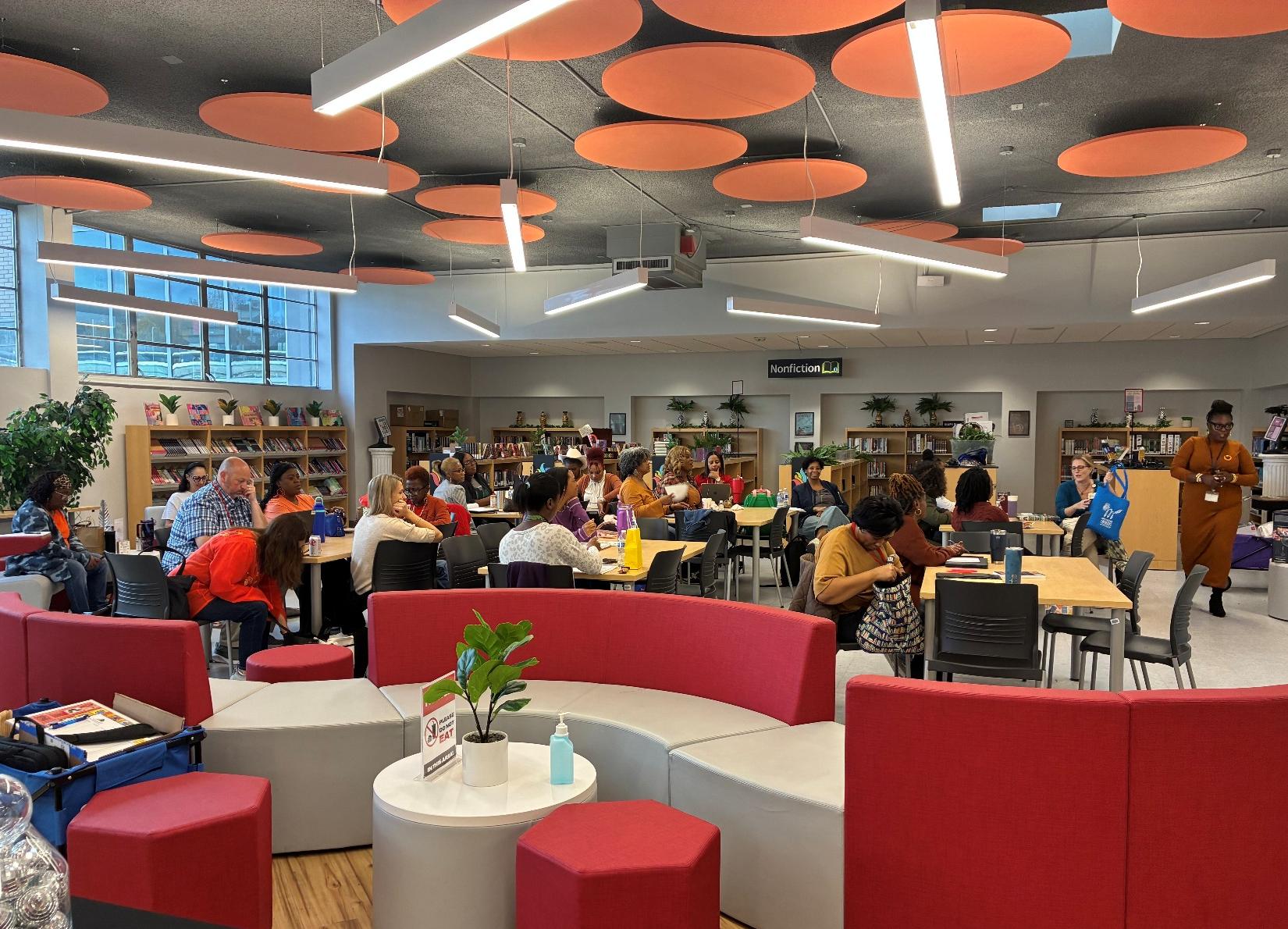 JPS Presenter A large group of people seated at tables in a modern library space with red and gray furniture.