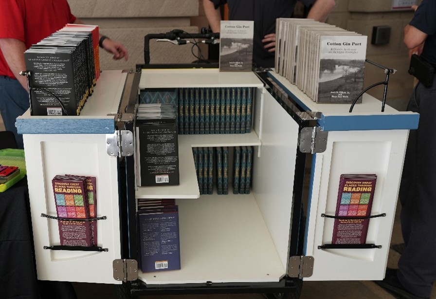 MLC-Book-Bike Open book bike display with shelves holding multiple books and pamphlets, including titles like ‘Cotton Gin Port’ and colorful reading guides.