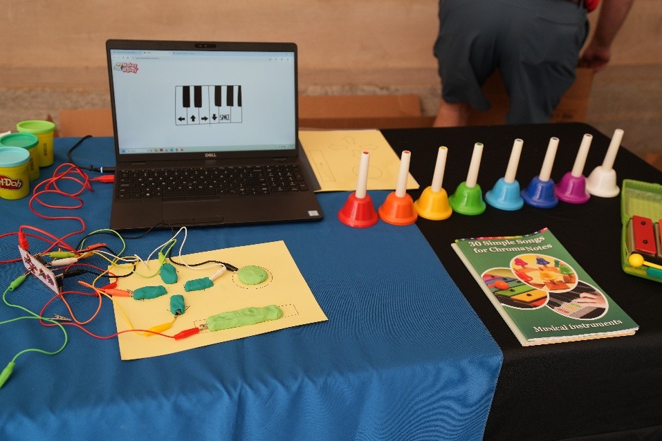 MLC-Games Table with a laptop showing piano keys, colorful handbells, Play-Doh containers, wires connected to a circuit board, and a booklet titled ‘36 Simple Songs for Chromaharp.’