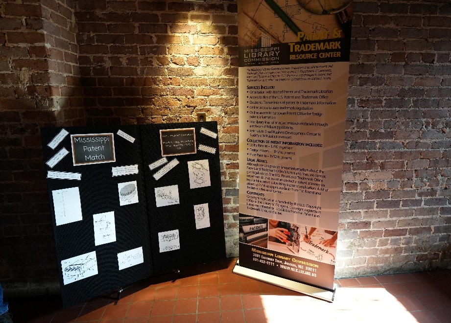 MLC-Posters Display featuring Mississippi Patent Match boards with sketches and a banner for the Patent and Trademark Resource Center against a brick wall.