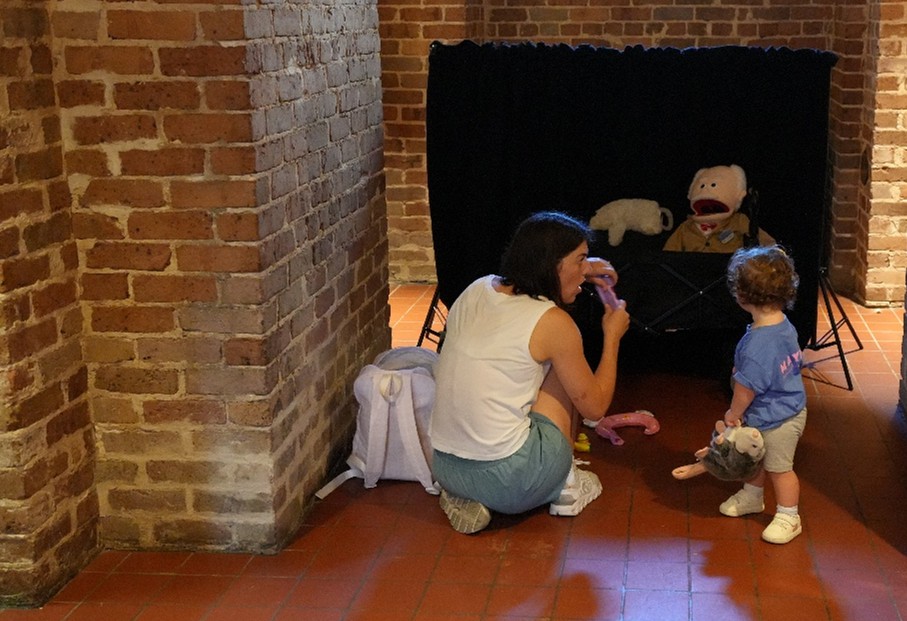 MLC-Puppets Child standing near a seated adult in front of a small puppet stage with stuffed animals and a large puppet inside a brick-walled room.