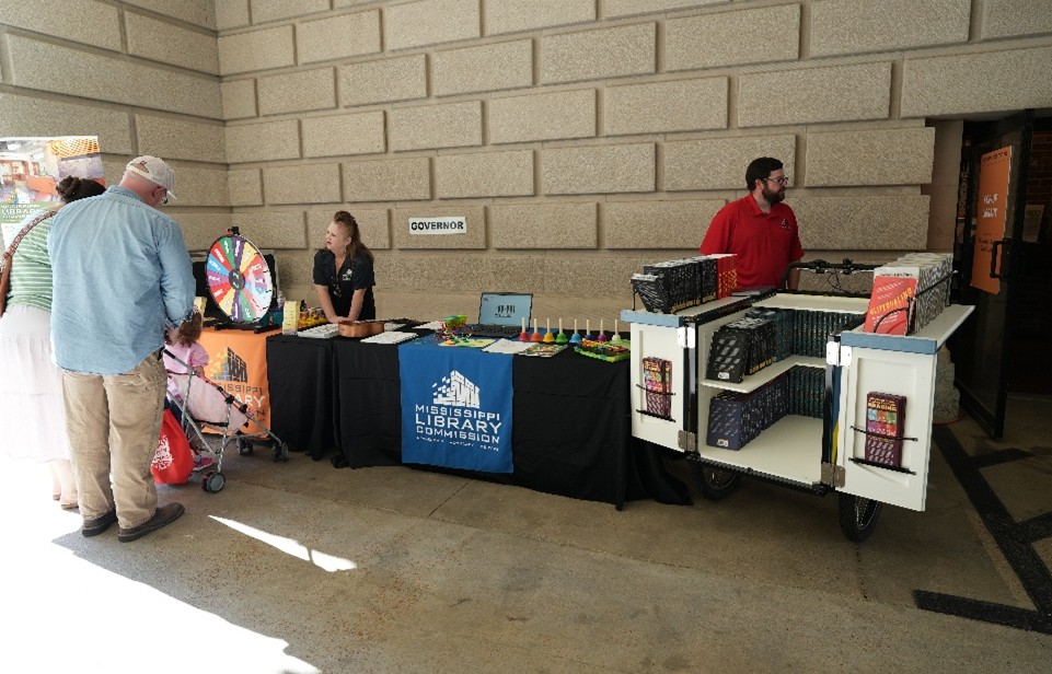 MLC-Special-Collections Mississippi Library Commission table with a prize wheel, colorful giveaways, and a cart filled with books in a hallway.