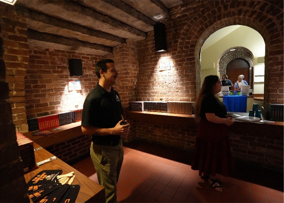 MLC-Staff Brick-walled room with wooden beams, books on shelves, and two people standing near a counter with a window opening to another area.