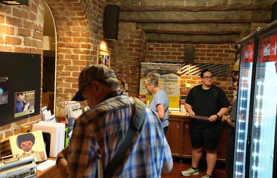 MLC-TBS Brick-walled room with informational displays and a banner for Talking Book Services, with people browsing materials on a wooden counter.