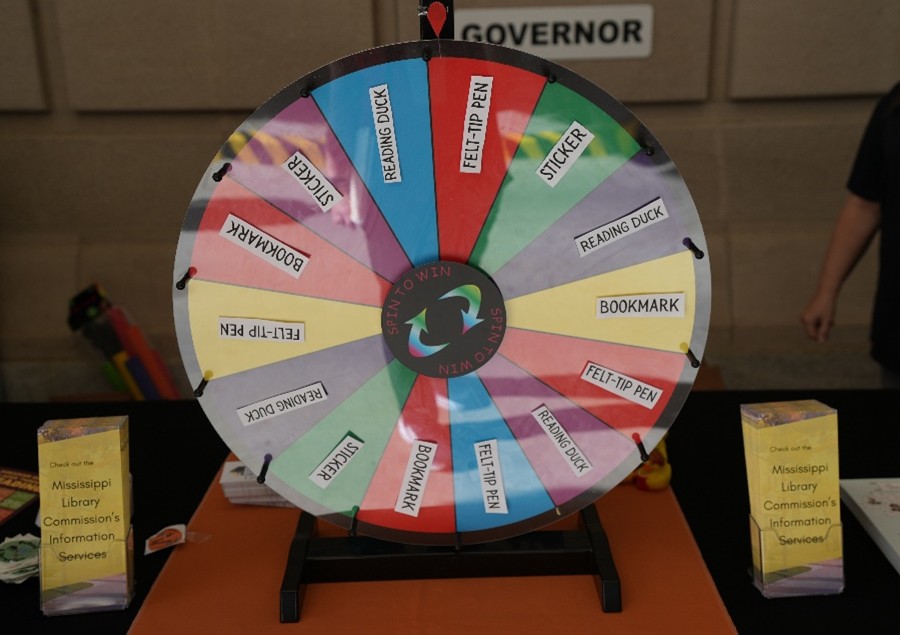 MLC-Wheel-Spin Colorful prize wheel labeled ‘Spin to Win’ with sections for items like stickers, bookmarks, reading ducks, and felt-tip pens, displayed on a table with brochures for Mississippi Library Commission’s Information Services.