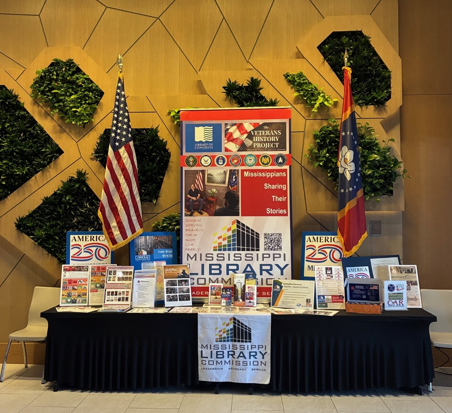 MML-01-Vendor-Display A Mississippi Library Commission display table is set up with brochures, books, banners, and two flags behind it. The center banner highlights the Veterans History Project and Mississippi library services.