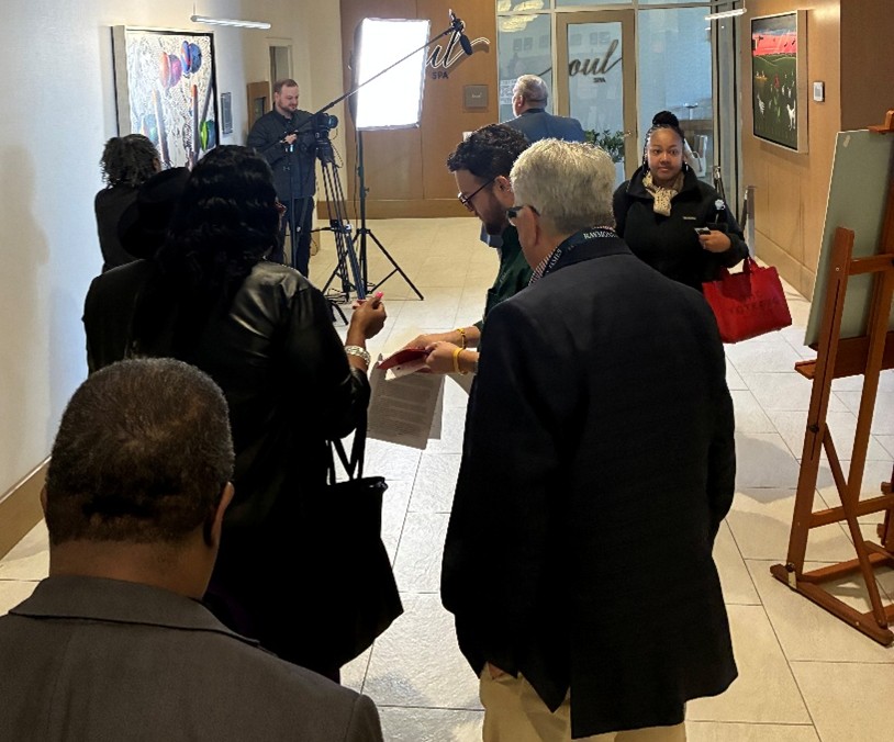 MML-02-Video-Recording-Line A group of attendees waits in a hallway near a video recording setup with lights and a camera operator. People are standing in line holding papers and moving toward the recording area.