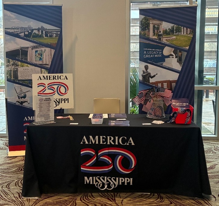 MML-03-AM250-Display A table featuring America 250 Mississippi promotional materials is displayed with banners and patriotic décor. Brochures, flyers, and small American flags are arranged neatly across the table.