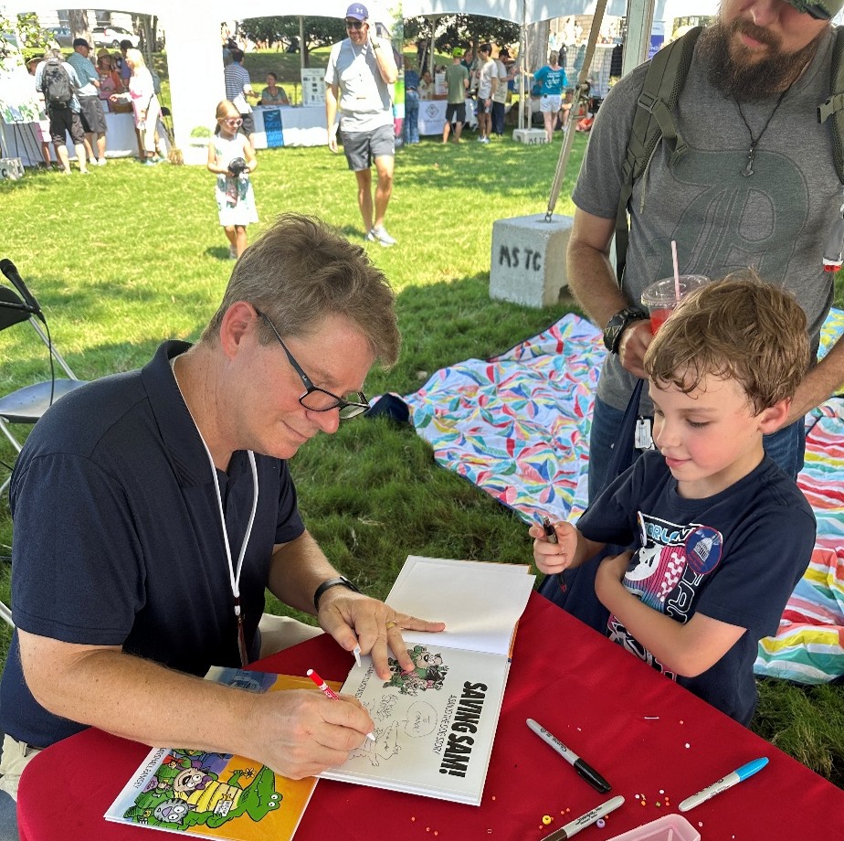 Marshall-Ramsey-Signing-Books Marshall Ramsey signing a book titled ‘Saving Sam’ at a table outdoors, with markers and a colorful blanket in the background.