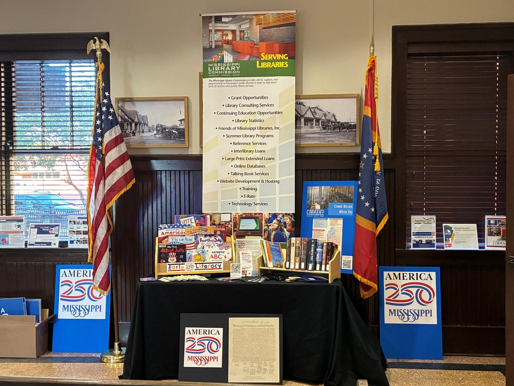 Natchez MLC Booth Mississippi Library Commission booth with books, America 250 posters, U.S. and state flags, and a banner listing library services.