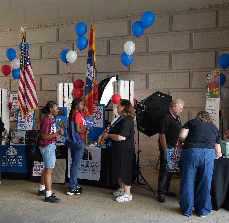People speaking at MS Book Fest People interacting at a Mississippi Library Commission booth decorated with balloons, flags, and America 250 posters.