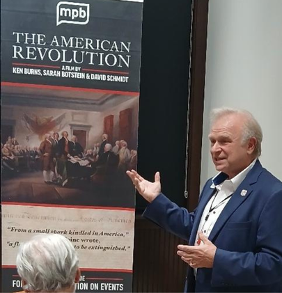 Speaking on the American Revolution Marvin Jeter gesturing toward a banner for the film ‘The American Revolution’ by Ken Burns, Sarah Botstein, and David Schmidt, featuring a historical painting and text about the event.