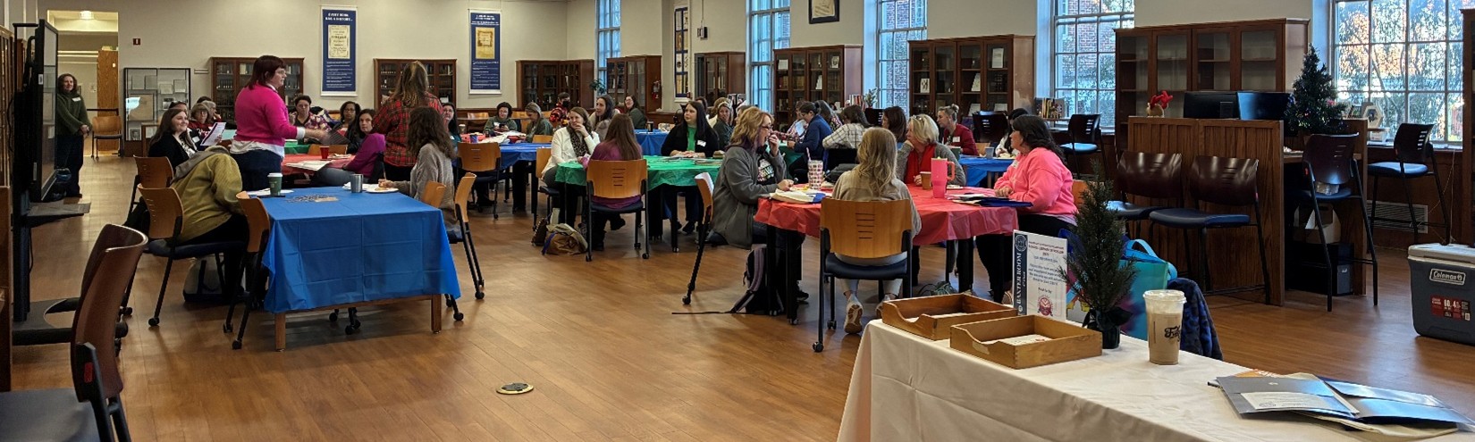 UM-Library-Symposium-01-Wide-Shot-Participants A large group of attendees sits at tables with colorful tablecloths during a session inside a library-style room. Two presenters stand at the front addressing the audience while bookshelves and large windows line the walls.