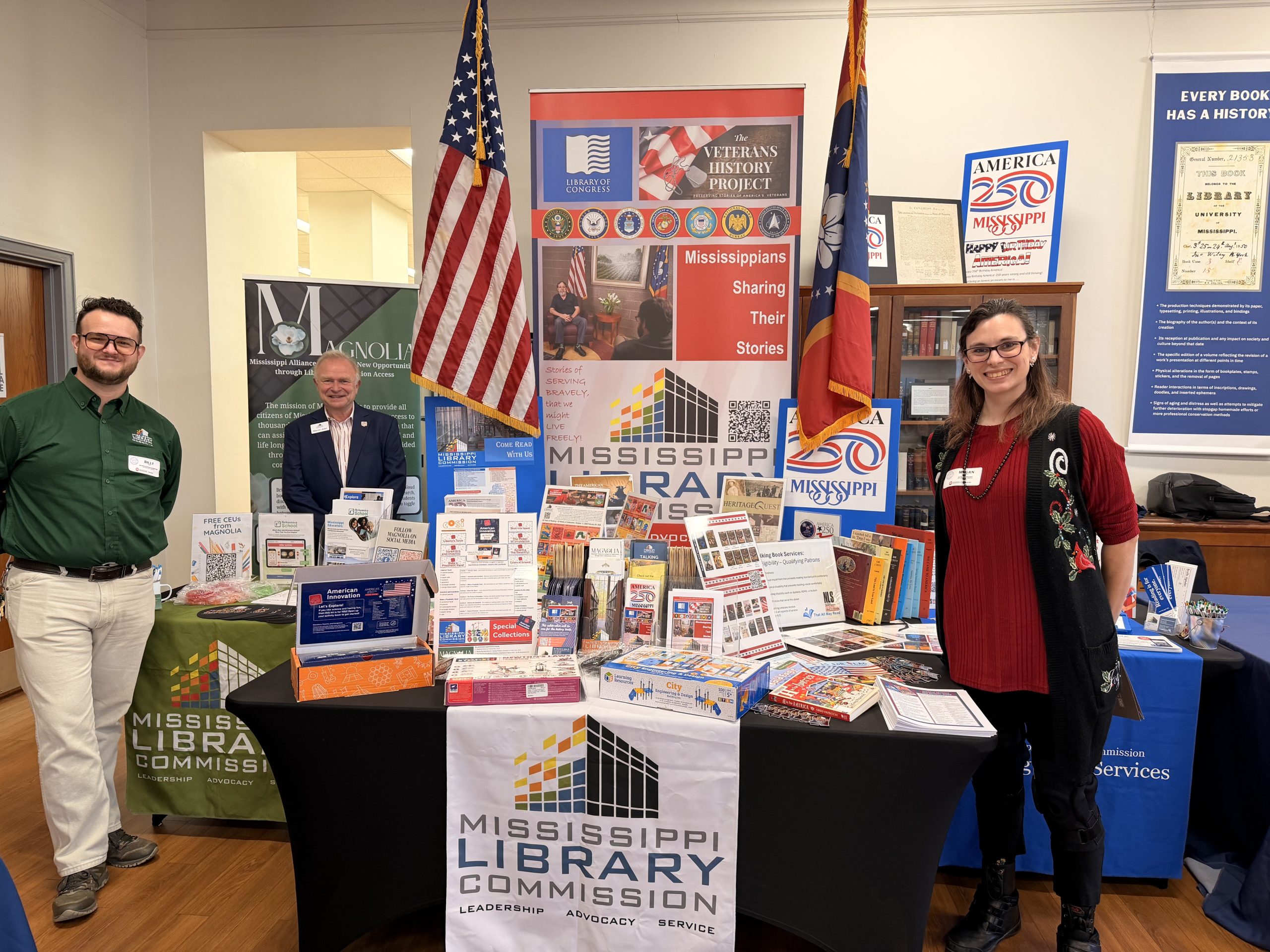 UM-Library-Symposium-03-Billy-Marvin-Megan-with-MLC-scaled Three people stand beside a large Mississippi Library Commission display table filled with books, flyers, and promotional materials. U.S. and state flags frame the colorful exhibit in the background.