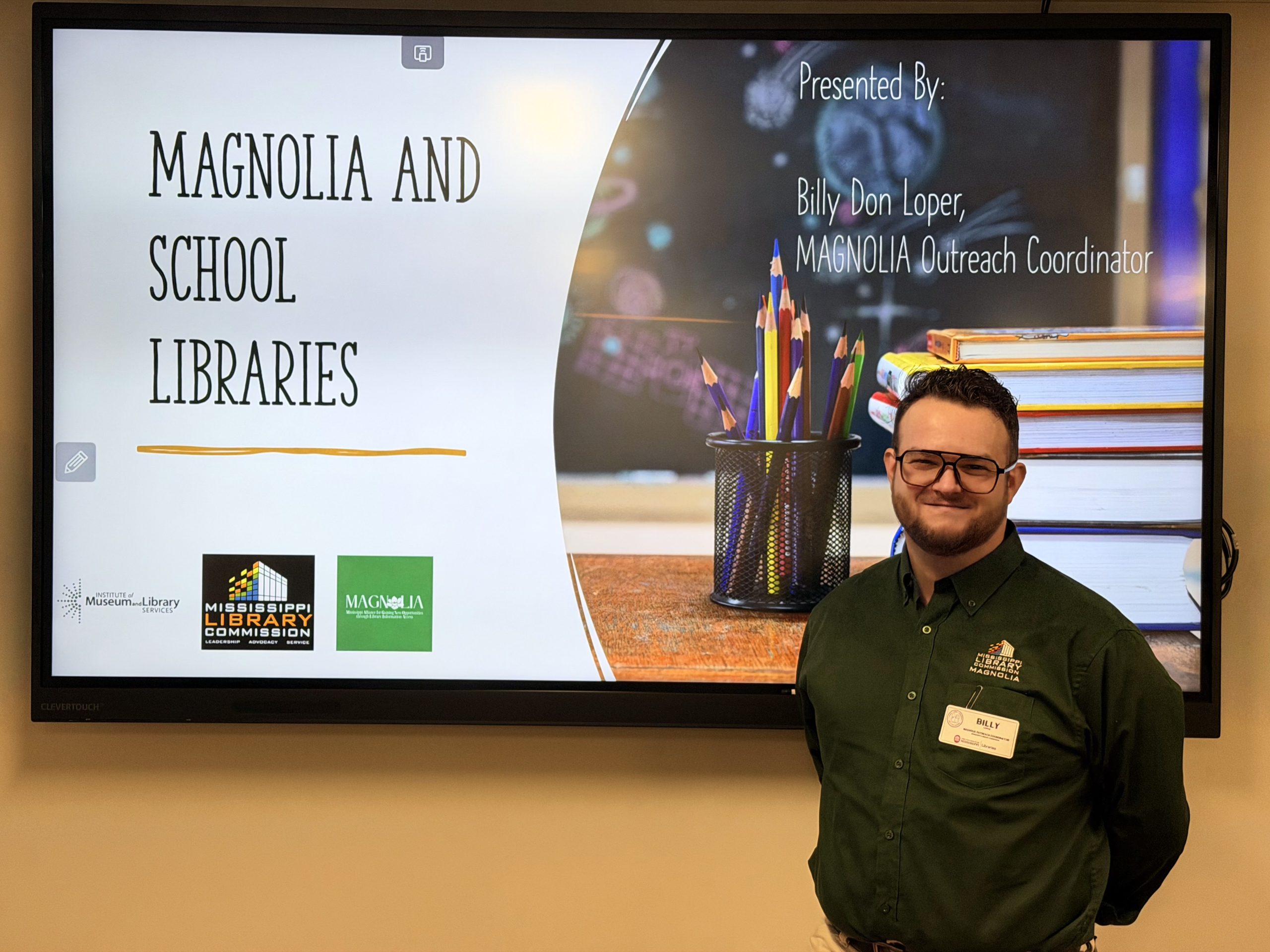 UM-Library-Symposium-07-Billy-Loper-scaled A presenter stands beside a large screen displaying a slide titled “Magnolia and School Libraries.” The slide shows school-themed graphics, including pencils and books, along with organizational logos.