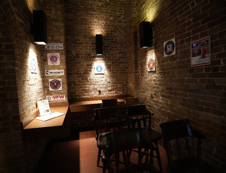 Veteran-History-Project-1 Brick-walled room with wooden chairs and tables, illuminated by wall lights, featuring posters for American Legion, VFW, and other veteran organizations.