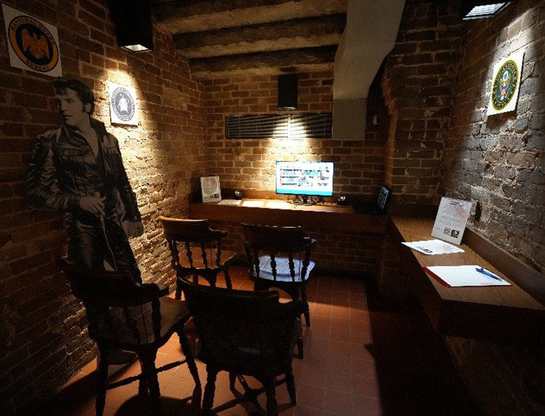 Veteran-History-Project-2 Brick-walled room with a computer on a wooden desk, chairs arranged in front, and posters for veteran organizations on the walls, with a cardboard cutout of Elvis.
